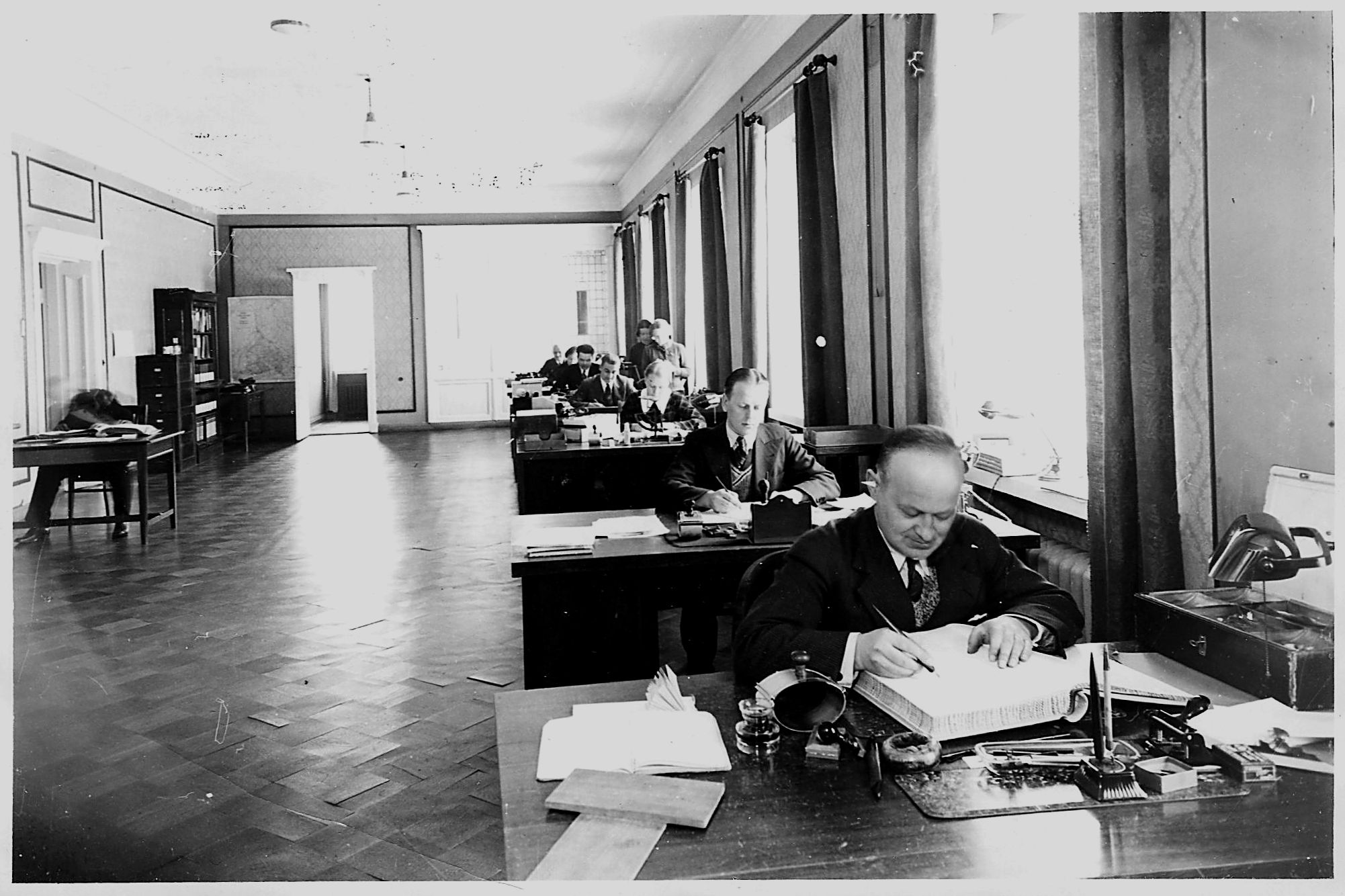 Konttorin sisällä on aina ollut paljon väkeä, joka ei ole paljasjalkaista. Tehtaankonttoristit istumassa oikealta: G.Relander, E,Carlson, Taimi Rautavaara (Kettunen), Bengt Enderlein, Bror Sjöstedt, Kerttu Relander (os.Wahlman), F.Michelsson. Vasemmalla tilintarkastaja Emil Malmberg liikahtaneena. Tunnetuin näistä lienee Georg Relander, kotiseutukirjailija Erik Relanderin isä, joka keräsi Kotiseutuyhdistykselle noin 3000 valokuvan pohjakokoelman äänekoskelaisista. Georg ja Kerttu asuivat Kotakennään sillanpielen yhä pystyssä olevassa Erland Ritasen rakentamassa talossa vuodesta 1935. Huomatkaa, että konttorityössä ei näkynyt vielä muita laitteita kuin paksut tilikirjat, mustepullo ja –kynä sekä kuivain. Merkillepantavaa on myös , ettei silmälaseja näytä kukaan vielä tarvitsevan! Ilmeisesti nykypäivänä koko tämä huoneellinen voitaisiin korvata yhdellä tietokoneen käyttäjällä ja kännykällä?